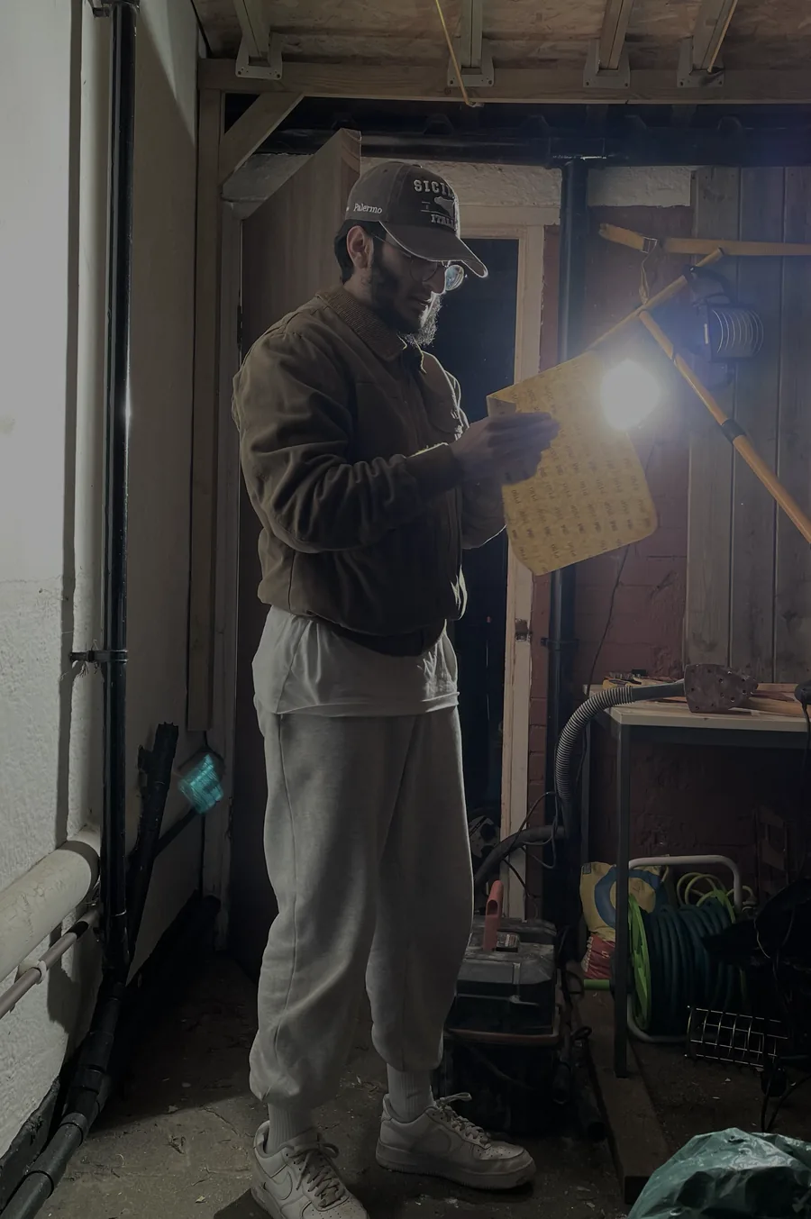 Ibrahim in a cap and jacket inspecting a sheet of sandpaper under a work light, with timber panelling behind him