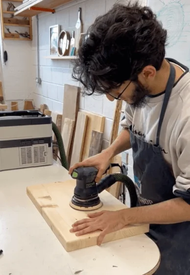 Ibrahim in a workshop apron sanding a pine board with an orbital sander